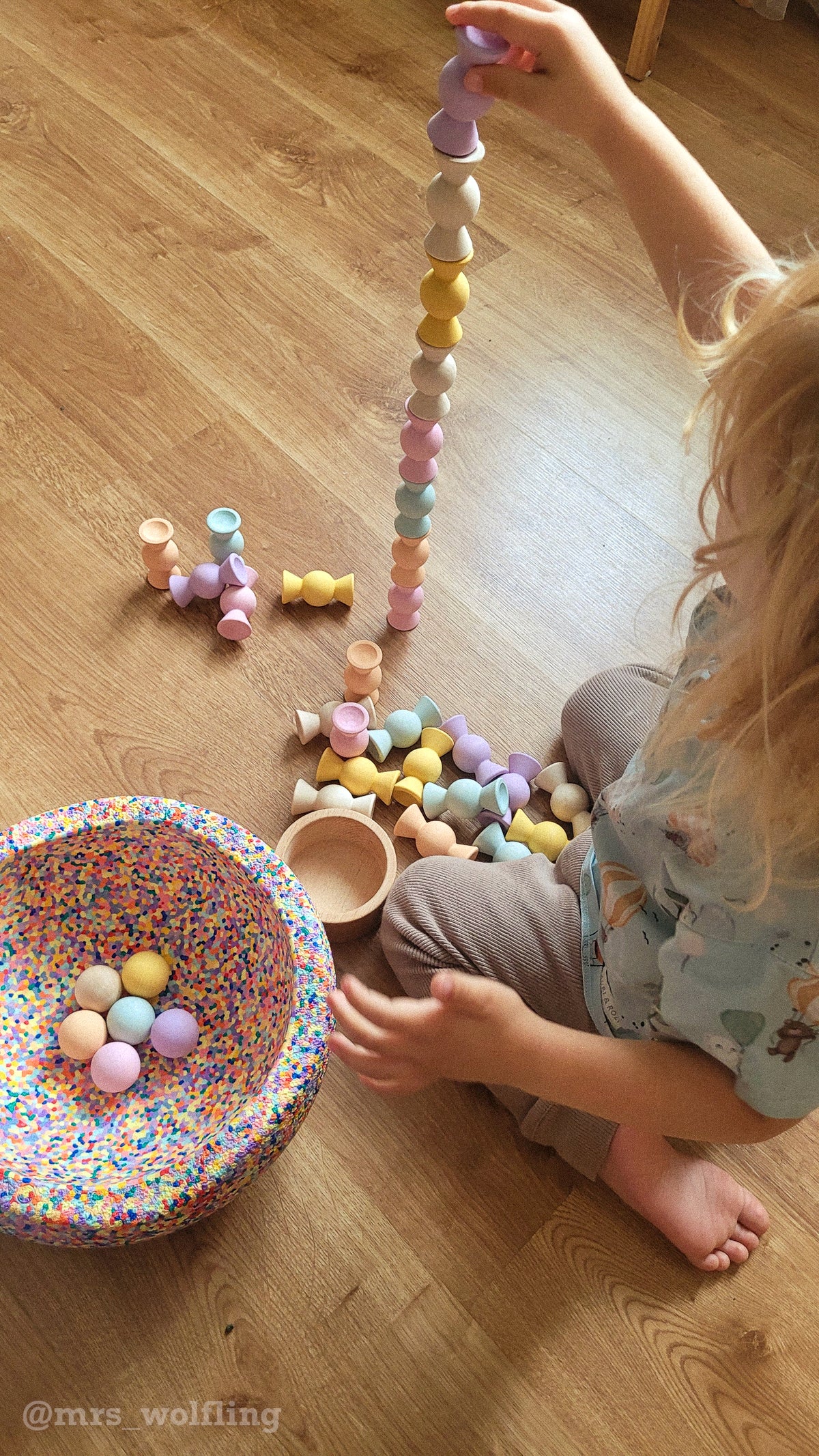 Child playing with pastel wooden pieces on the floor beside a tall tower and a circular arrangement of toys.