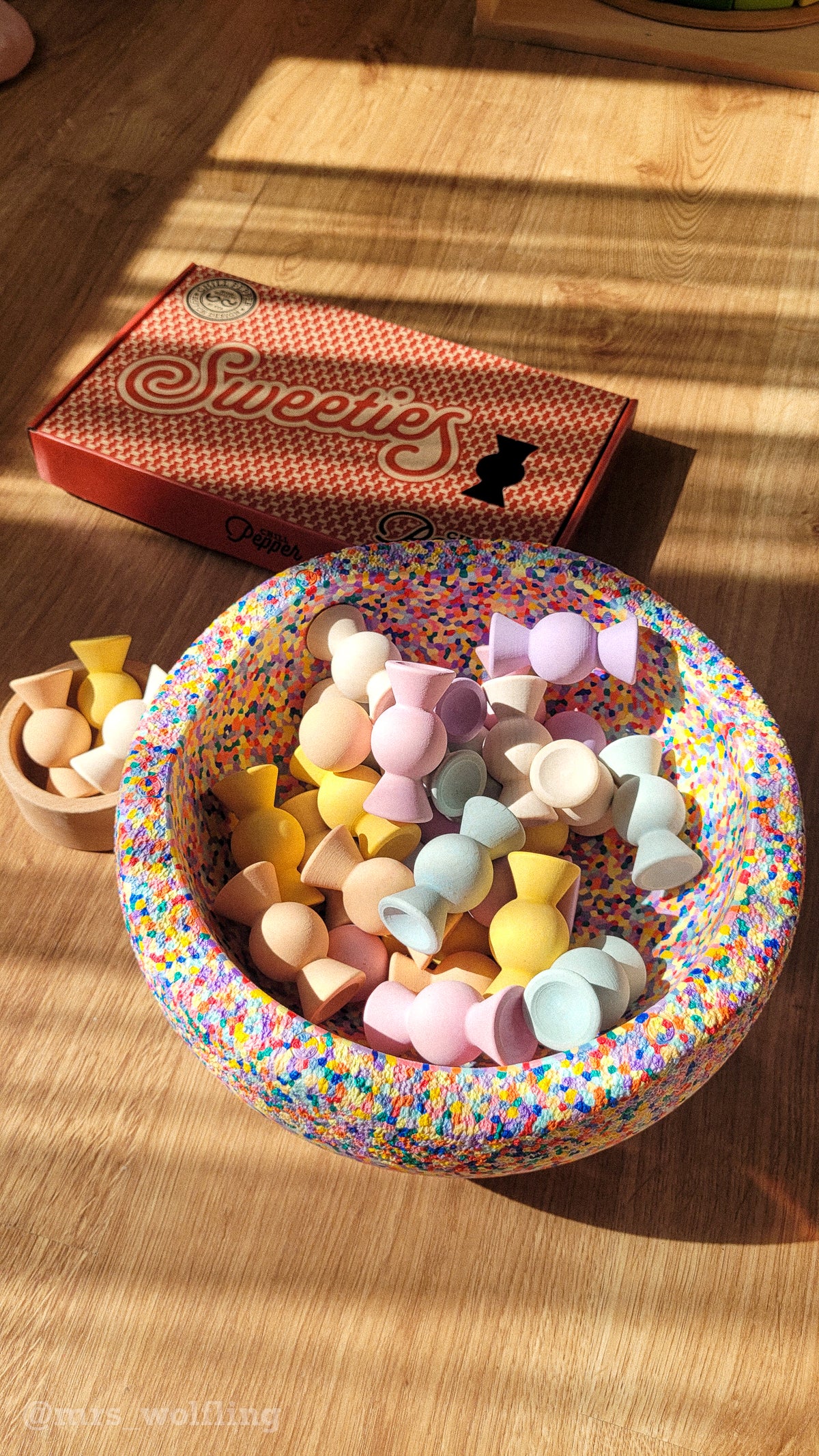 Overhead view of Chill Pepper Sweets Peachy Pastels wooden toys in a colourful speckled bowl beside their patterned box on a sunlit wooden floor.